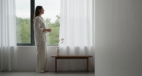 زنی ایستاده کنار پنجره دارای پرده های سفید از نمای دور داخل خانه