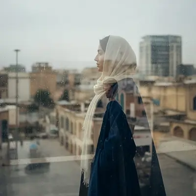 زن با پوشش ساده و شال روشن، ایستاده پشت شیشه در روز بارانی و نگاه‌کردن به منظره شهری؛ حس مکث، تنهایی و تأمل در زندگی شهری.